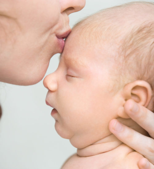 Funny newborn babe napping in mom arms. Young mother kissing sleeping adorable new born child on the forehead. Healthy little kid in parent arms. Love, bonding, happy family concept. Indoor. Close-up